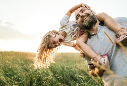 Eine Person trägt ein Kind auf den Schultern in einer grünen Wiese bei Sonnenuntergang.